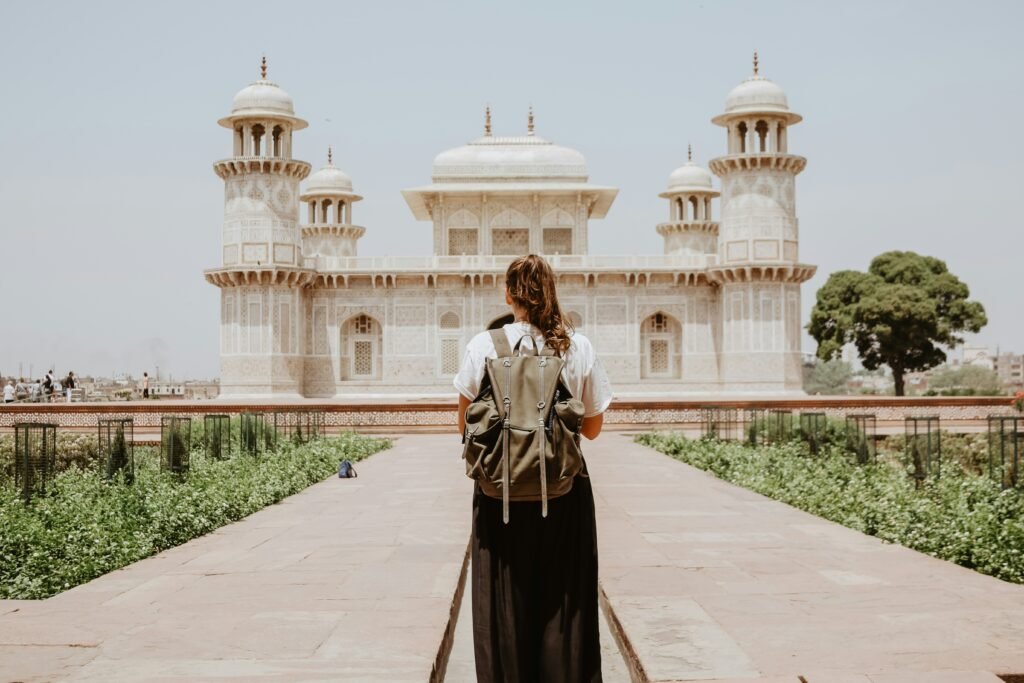 A person with a backpack stands on a path facing an ornate white stone building with domes and towers, surrounded by gardens.